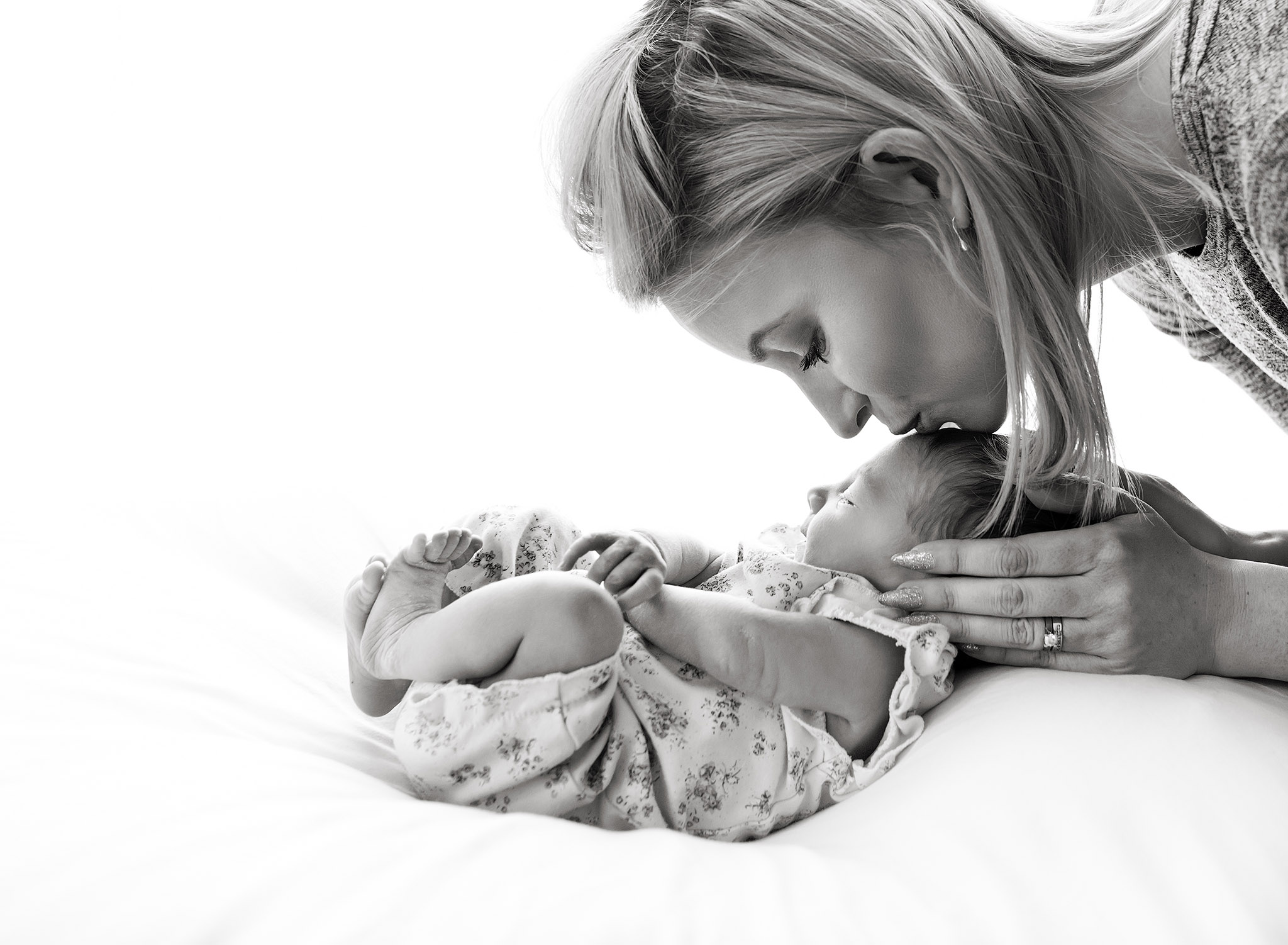 black and white image of a mother kissing her newborn baby on the forehead, lying on a white cushion at Fairy Nuff Photography, Nottingham