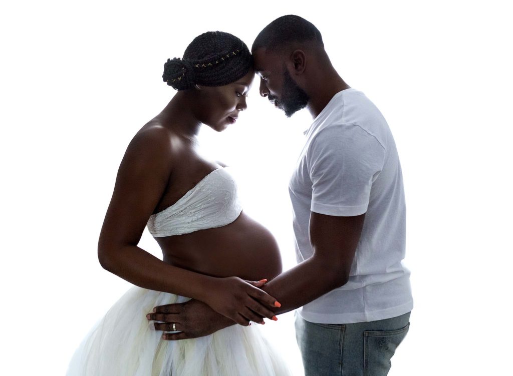Pregnant couple, dressed in white against a white backdrop facing each other