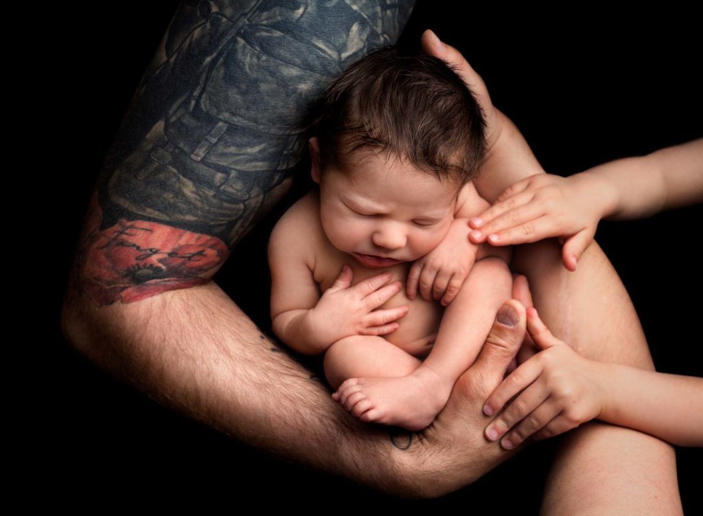 Newborn baby cradled in his parents and sibling arms against a black background at a baby photoshoot