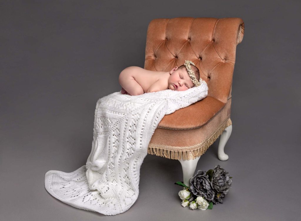Baby girl posed on an antique chair with flowers and a white christening blanket a a baby photoshoot