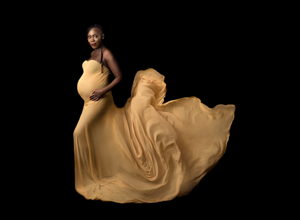 Pregnant woman standing sideways in a yellow flowing dress, holding her bump I a maternity photo session.