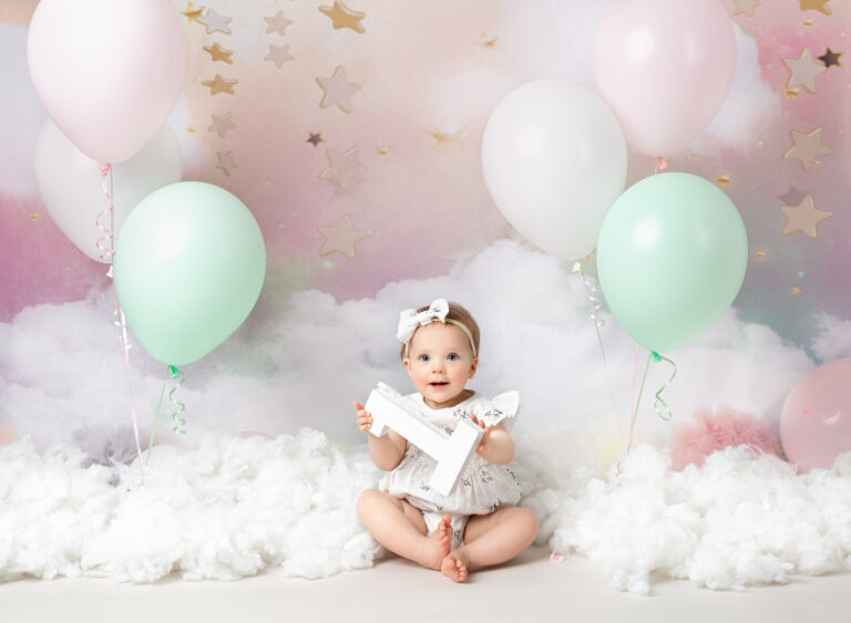 1st Birthday set with baby girl in white romper, holding white number 1 in her hands against a pink cloud backdrop 1st Birthday photography session, Fairy Nuff Photography, Nottingham