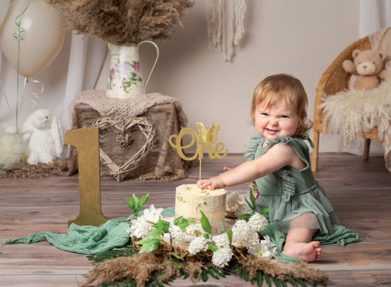 Boho theme cake smash session for a one year old. Baby girl in a green outfit smiling and putting her hands in the cream birthday cake. 1st Birthday photography session, Fairy Nuff Photography, Nottingham