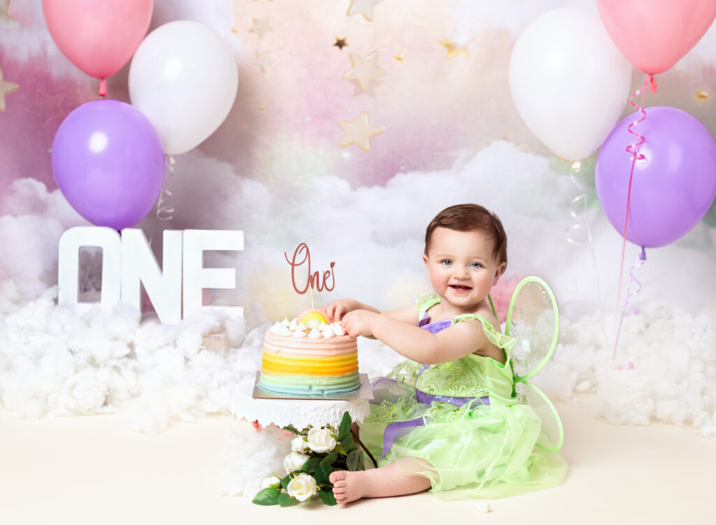 1st Birthday cake smash photoshoot with Fairy Nuff Photography Nottingham. Baby girl in a green fairy outfit with hands in her rainbow cake. Rainbow backdrop and balloons.