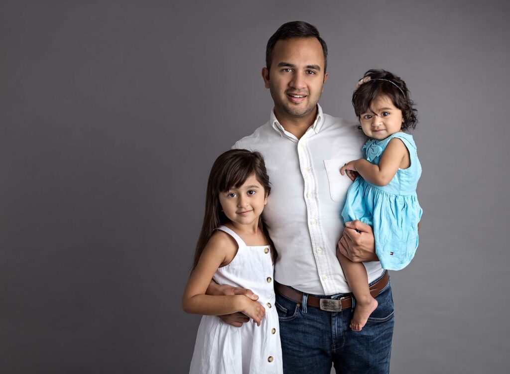 Dad holding Daughter with older Daughter standing next to him against a grey background at a family photoshoot with Fairy Nuff photography, Nottingham