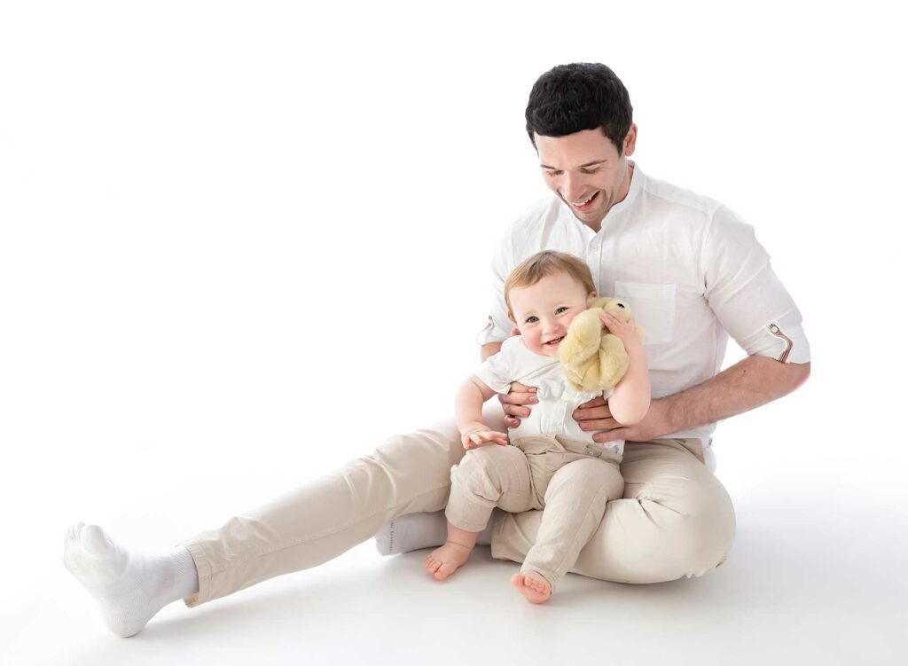 Dad and baby son dressed in cream and white with a white background seated on the floor. Family photoshoot at fairy Nuff photography, Nottingham